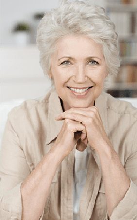 Head-and-shoulders photo of 65+ woman with gray hair sitting on a white couch and smiling with her chin resting on her hands - for information on implant overdentures from Kentucky Dental Group of Lexington.